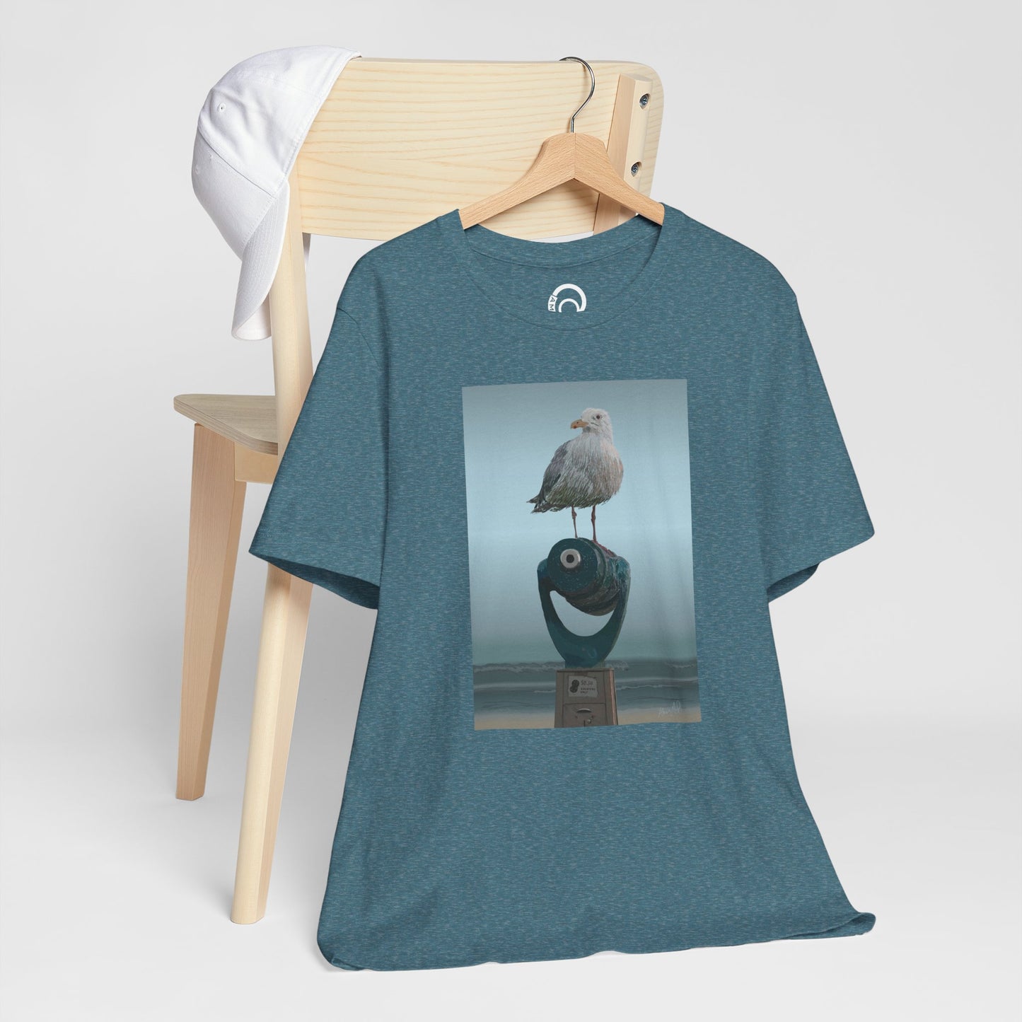 Heather Deep Teal Tee, hanging ion the side of a wooden chair, with a white baseball cap