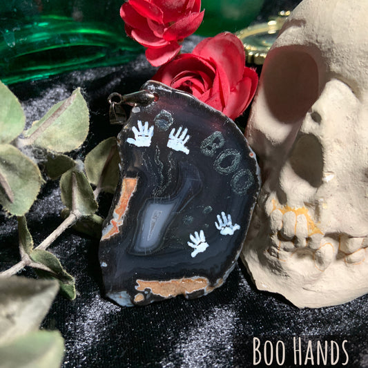 Four white hand prints are hand-painted on a dark grey agate slice, with a white group of crystals in the lower-left side. Two patches of orange stone interrupt the smooth surface.