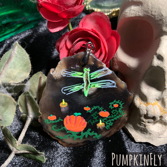 A hand-painted green dragonfly hovers over a pumpkin patch lit with candles. Within the dragonfly's body are naturally formed crystals inside the brown agate slice.